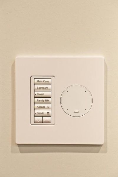 Wall-mounted control panel with multiple labeled buttons for different room lights and shades.