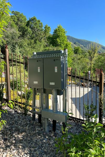 Outdoor electrical control boxes installed on metal stands, surrounded by greenery and a scenic mountain view in the background.