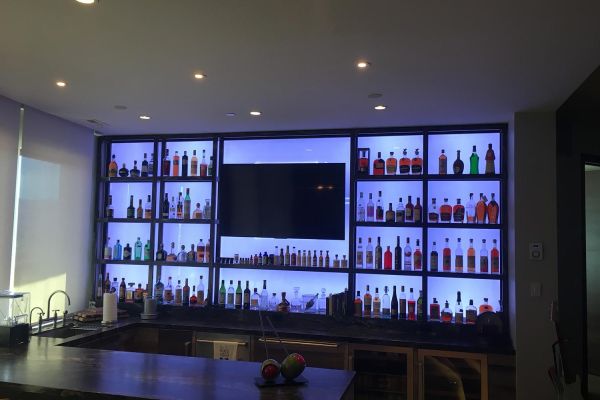 A modern home bar with backlit shelves displaying an extensive collection of bottles, centered around a large television.