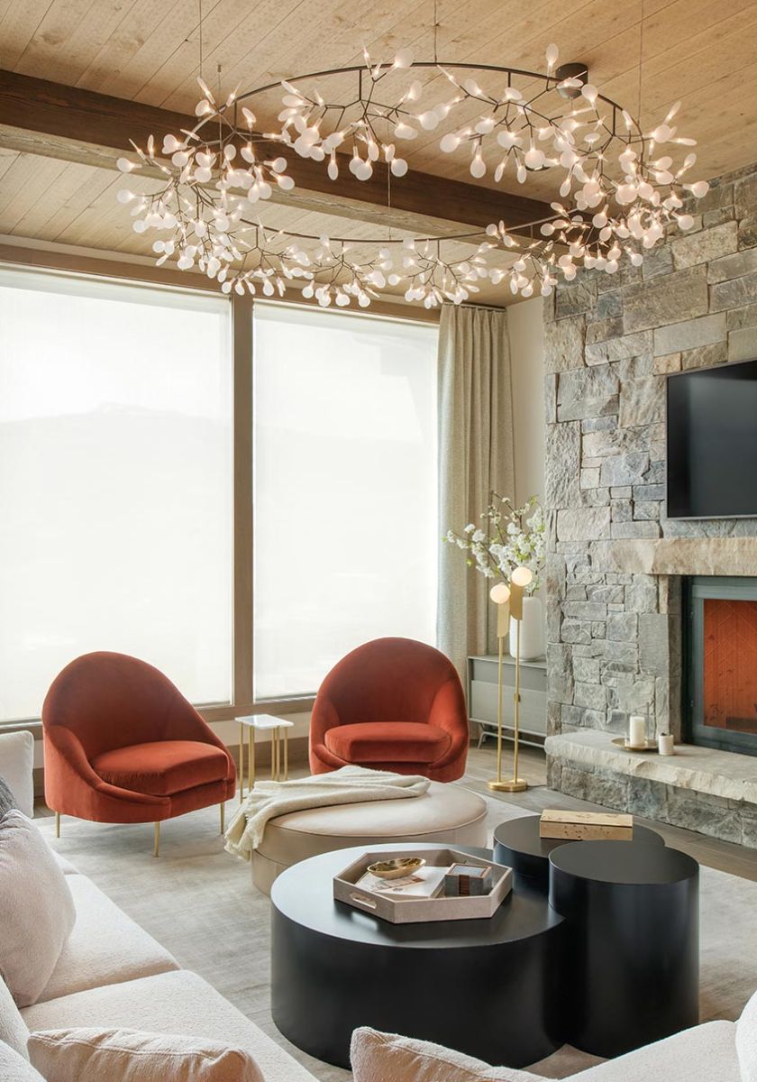 A cozy living room featuring a stone fireplace, two red armchairs, a large circular chandelier with a delicate branch design, and light filtering through tall windows, enhancing the warm and inviting space.