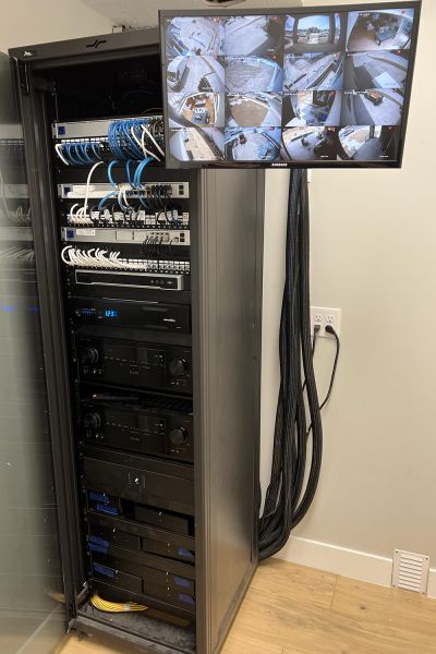 A network rack with neatly organized cables and equipment, next to a wall-mounted security monitoring screen.