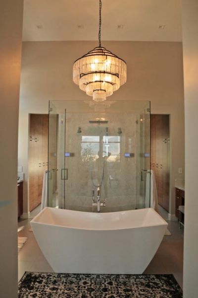 An elegant chandelier with tiered, translucent shades illuminates a luxurious bathroom with a central freestanding bathtub and glass shower enclosure.