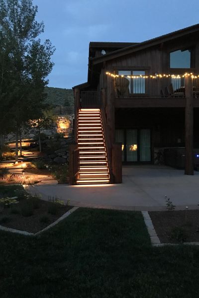 Wooden deck with illuminated stairs leading to a second-story entrance, surrounded by garden lights.