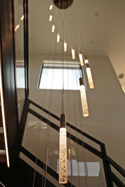 A modern, cascading light fixture with multiple slender, cylindrical pendants hanging in a vertical arrangement in a stairwell.