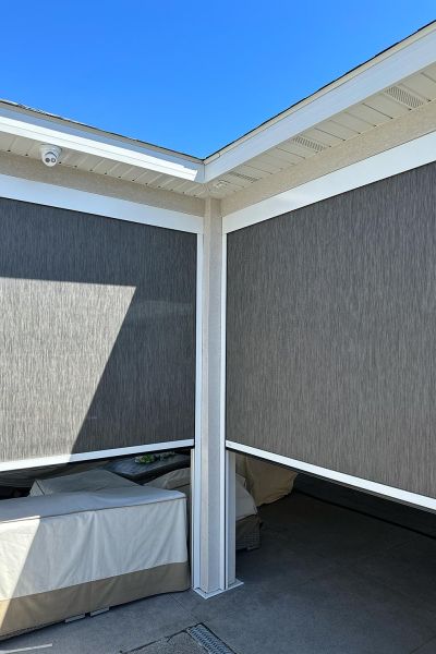 An outdoor seating area with motorized retractable screens partially lowered, creating a shaded, private space under a clear blue sky.