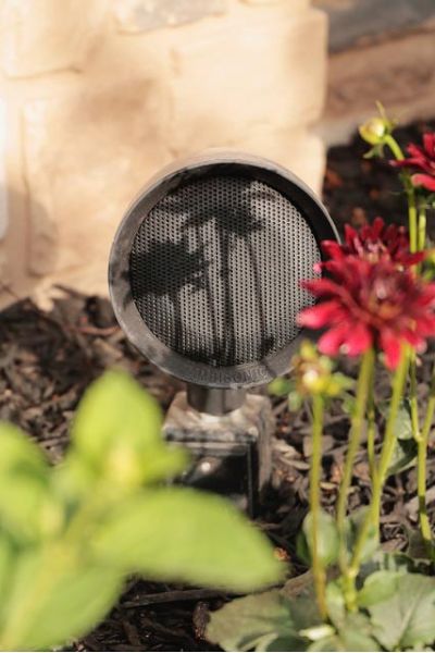 Outdoor garden speaker installed among red flowers and mulch, with a stone wall in the background.