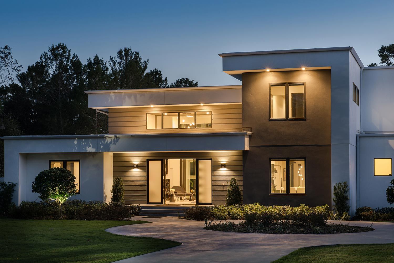 A modern two-story house with exterior lighting, large windows, and a landscaped front yard, photographed at dusk.