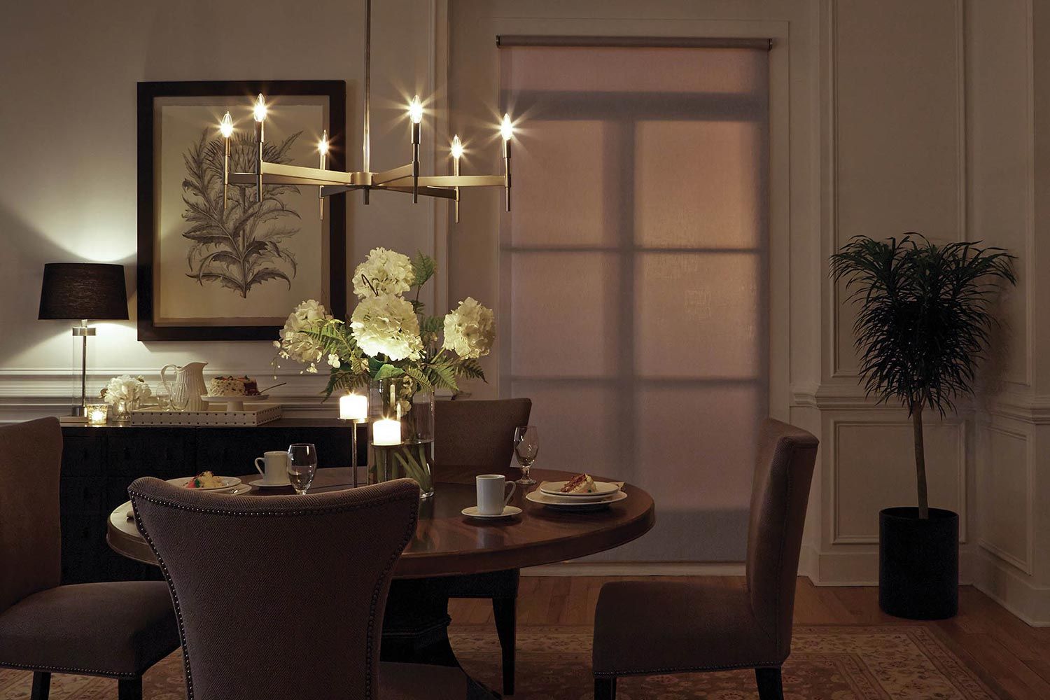 A dining room with a round table, chairs, a modern chandelier, flowers on the table, and a large framed artwork on the wall.