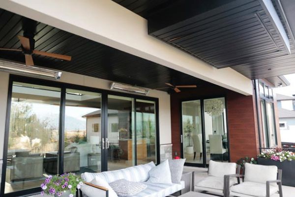 Covered patio with seating area, ceiling fans, and outdoor heaters, overlooking a modern home interior through glass doors.