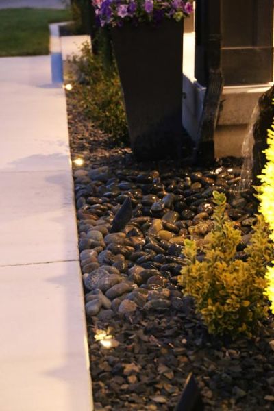 Evening view of a landscaped area with small pathway lights, decorative stones, and plants near a building's corner.