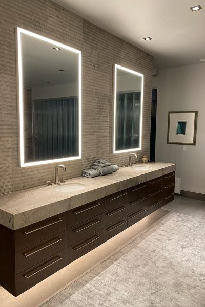 A contemporary bathroom with a long vanity, illuminated mirrors, and under-cabinet lighting, providing a sleek and clean appearance.