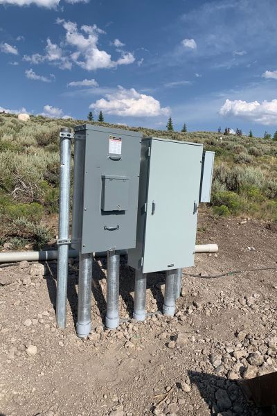 Outdoor electrical control panel with multiple gray boxes and conduit pipes in a rural setting.