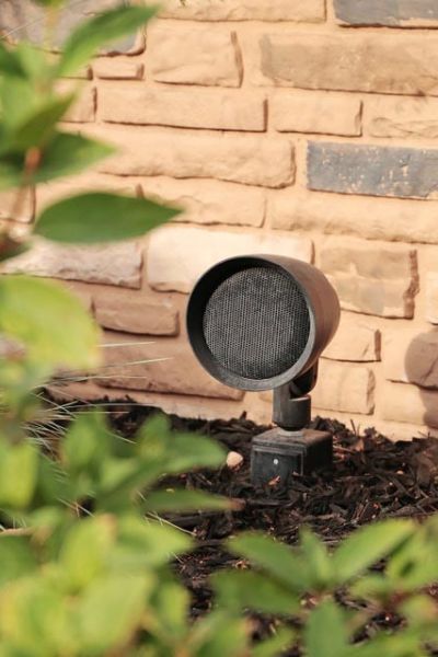 Close-up of a small outdoor speaker installed in a landscaped garden with a brick wall in the background.