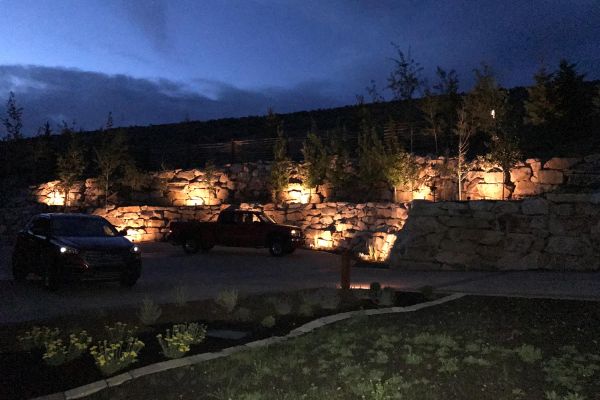 Nighttime scene with cars parked in front of a landscaped retaining wall with accent lighting.