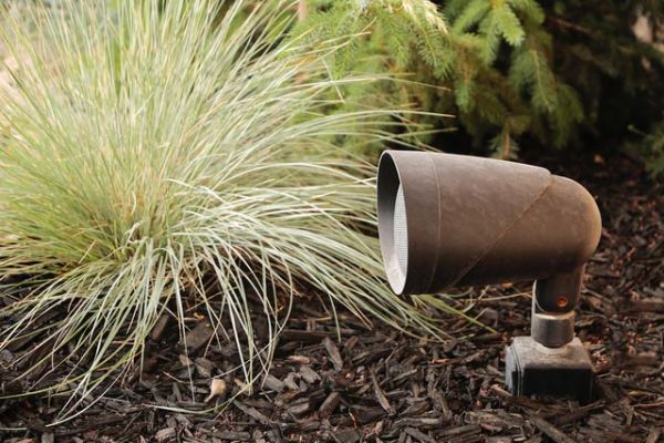 Outdoor landscape speaker installed in a garden bed, surrounded by mulch and ornamental grasses.