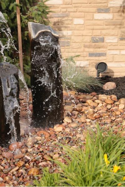 Outdoor water feature with two vertical stone fountains surrounded by river rocks in a landscaped garden.